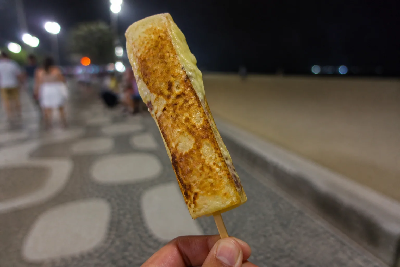 Crepe no palito dourado, servido no palito de madeira, segurado na mão numa noite de calçadão em Capão da Canoa